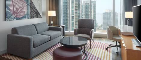 A modern hotel room with a grey sofa, a round coffee table, a red ottoman, a TV, and a large window offering a city view.
