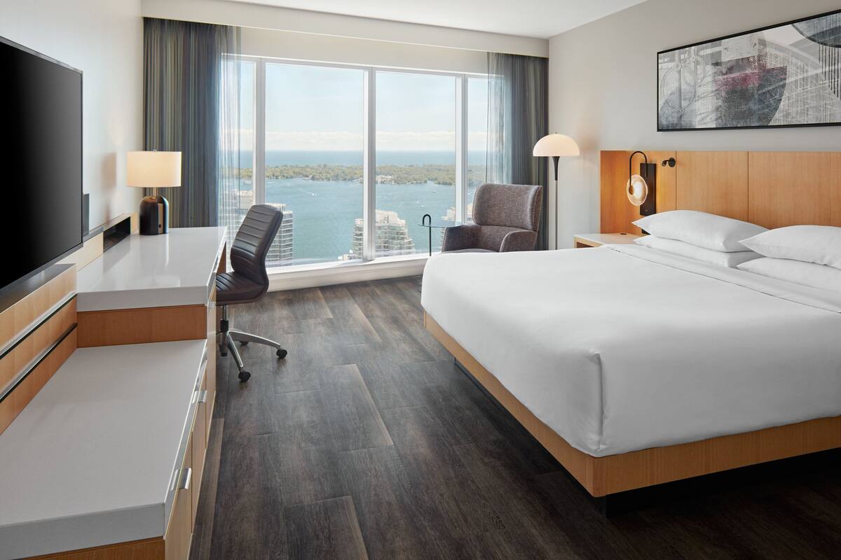 Quarto de hotel com uma cama grande, uma mesa com cadeira, televisão e vista da cidade por grandes janelas.