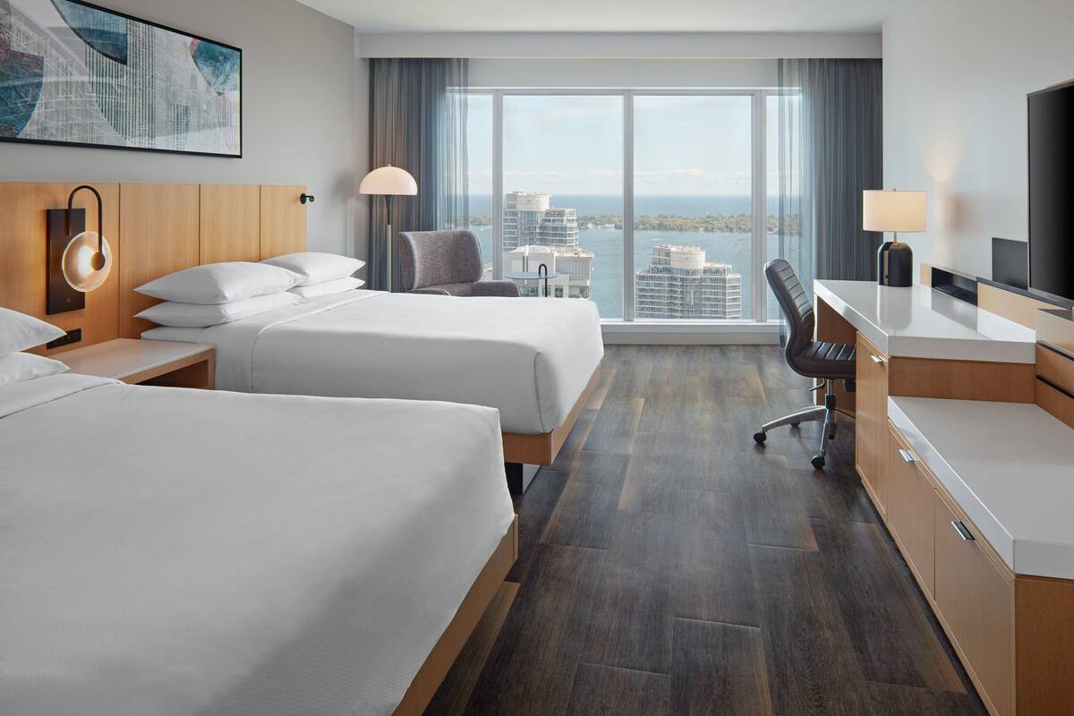 Quarto de hotel com duas camas, uma escrivaninha, uma cadeira, uma televisão e uma janela grande com vista para a cidade.