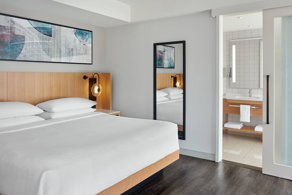 Quarto de hotel moderno com uma cama grande, cabeceira de madeira, mesa de cabeceira com luminária, espelho amplo e vista para o banheiro com pia e porta-toalhas.