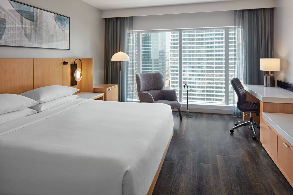 Quarto de hotel com uma cama grande, uma mesa, uma cadeira e vista para edifícios altos pela janela.