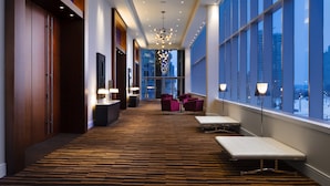 Un pasillo de hotel con alfombra estampada, zonas de estar y grandes ventanales con vistas a la ciudad.