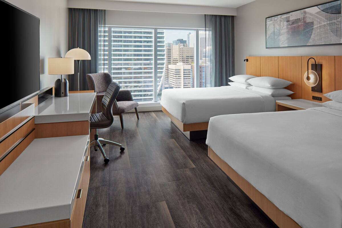 Quarto de hotel com uma cama grande, uma cama menor, uma escrivaninha com abajur, uma cadeira e uma televisão.