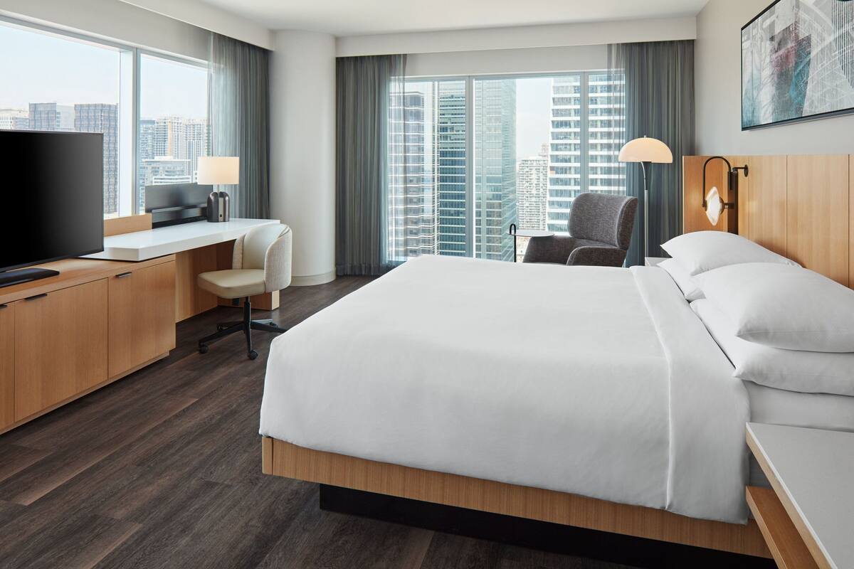 Quarto de hotel com uma cama grande, uma escrivaninha com televisão, uma cadeira e vista para prédios altos.