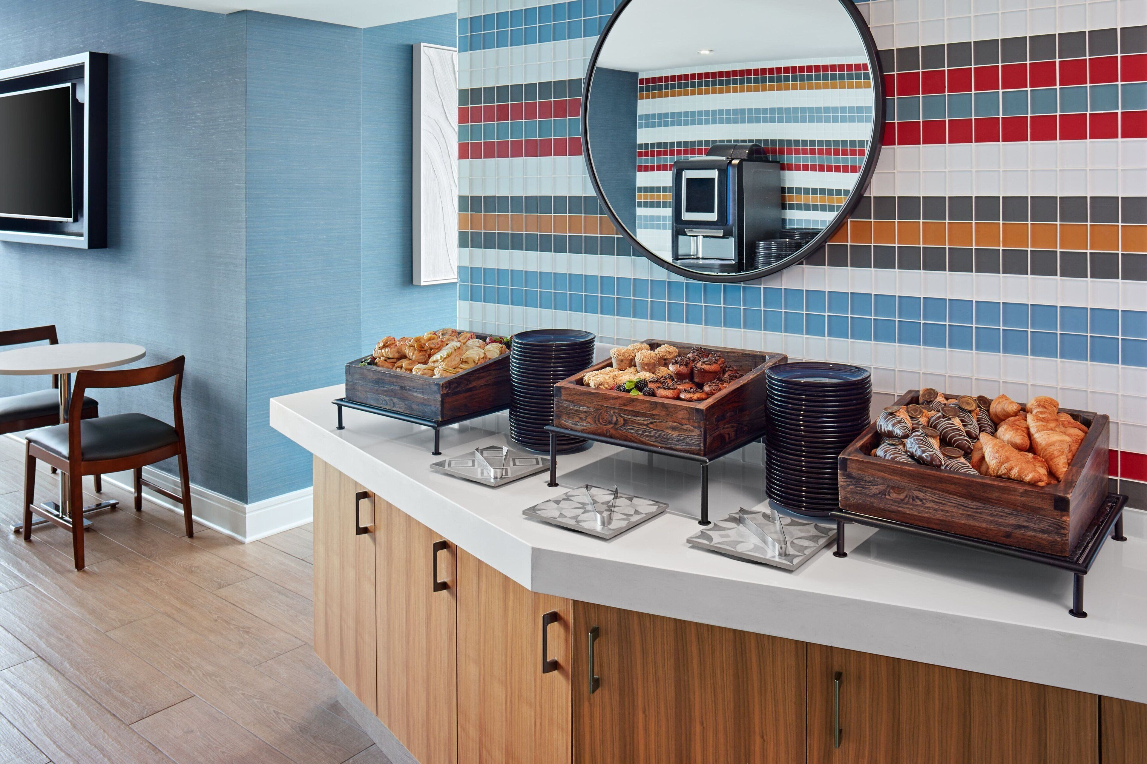 Un bufé de desayuno con diversos alimentos, un televisor montado en la pared y un espejo que refleja la habitación.
