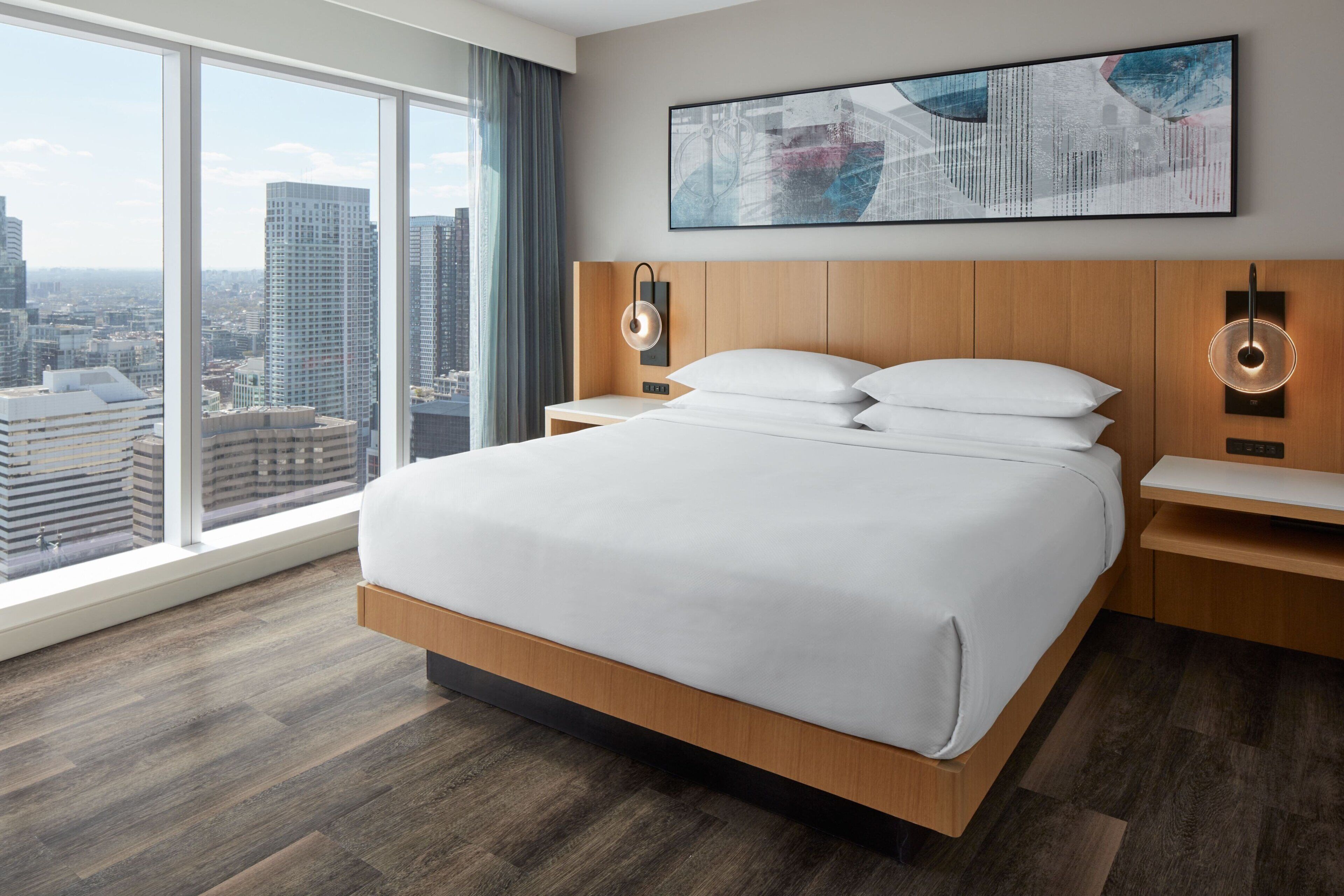 Une chambre d’hôtel moderne dotée d’un grand lit, d’une tête de lit en bois et offrant une vue sur la ville grâce à de grandes fenêtres du sol au plafond.