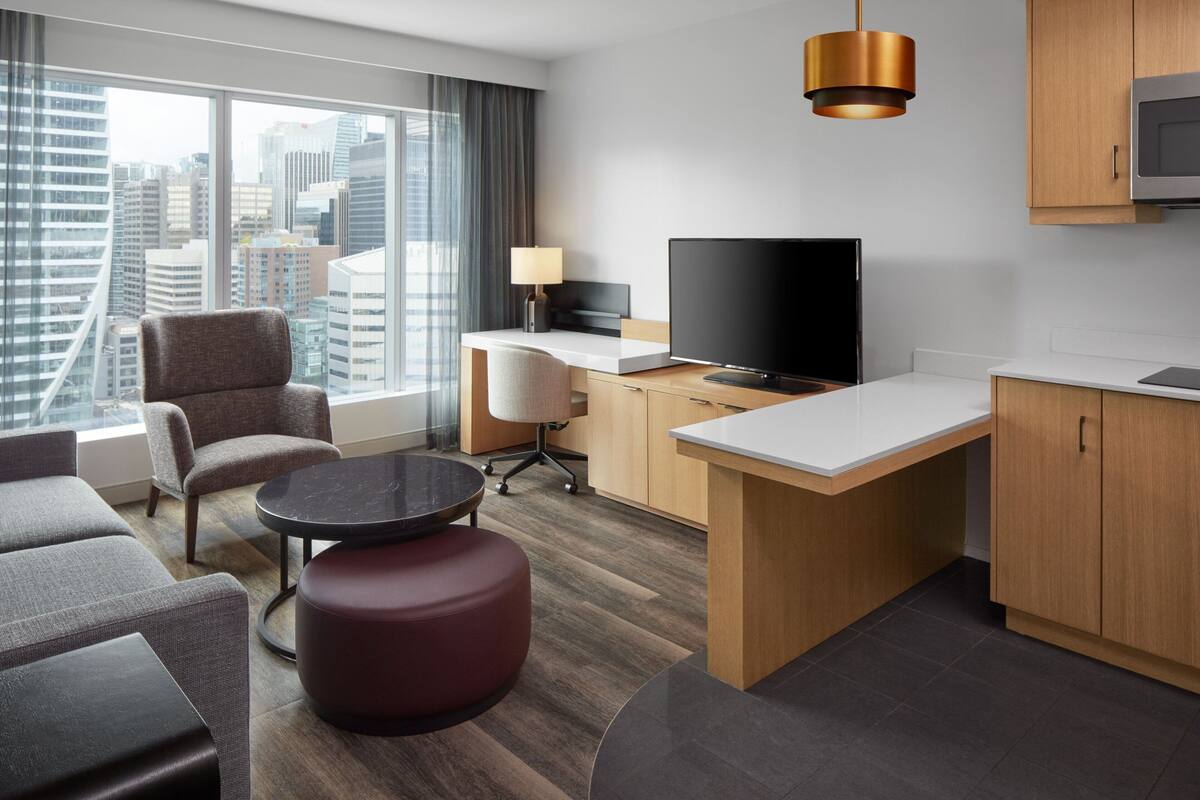 Quarto de hotel moderno com uma janela grande com vista para a cidade, uma TV de tela plana, uma área de refeições e um espaço de estar com um banquinho vermelho.