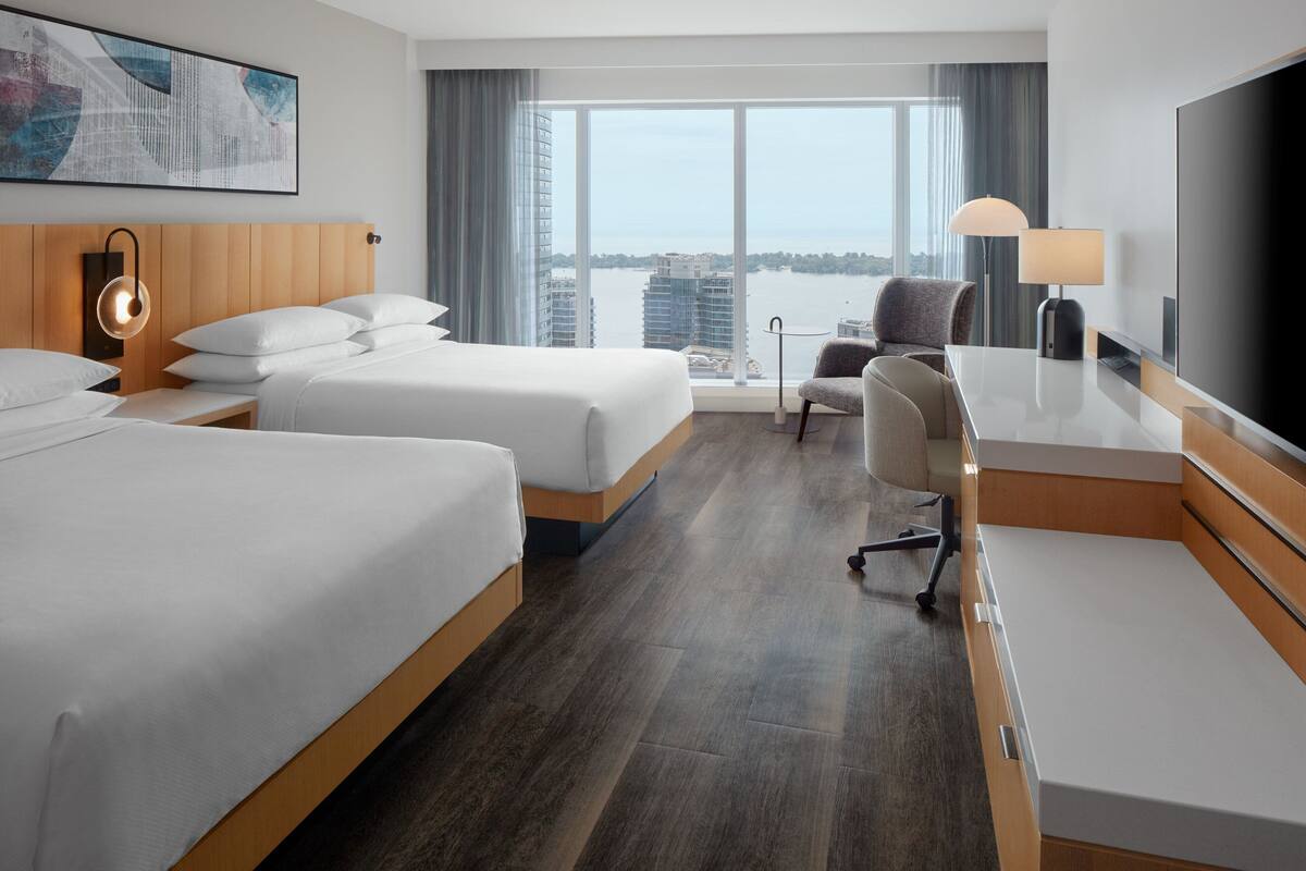 Quarto de hotel com duas camas, uma escrivaninha, uma cadeira, televisão e uma janela ampla com vista para a cidade.