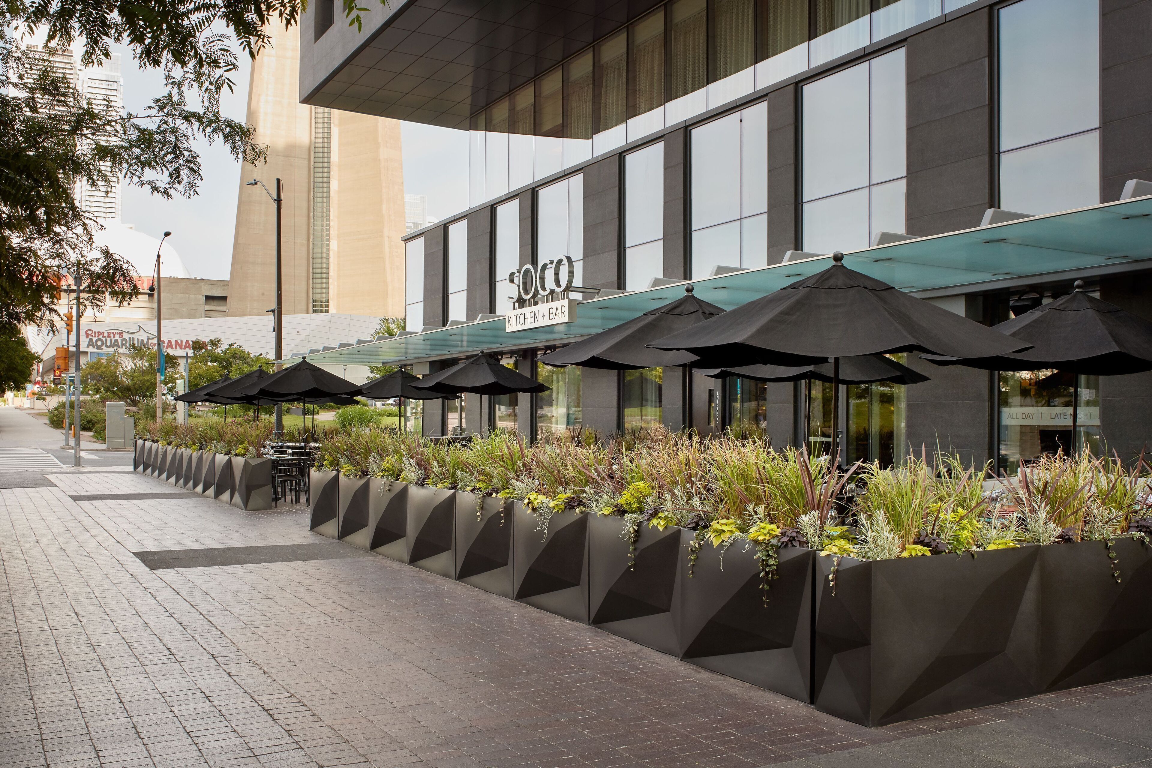 A modern building with a restaurant named "Soco Kitchen + Bar" featuring outdoor seating with umbrellas and planter boxes with plants.