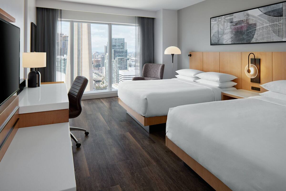 Quarto de hotel com duas camas, uma escrivaninha, uma cadeira, televisão e uma janela grande com vista para a cidade.