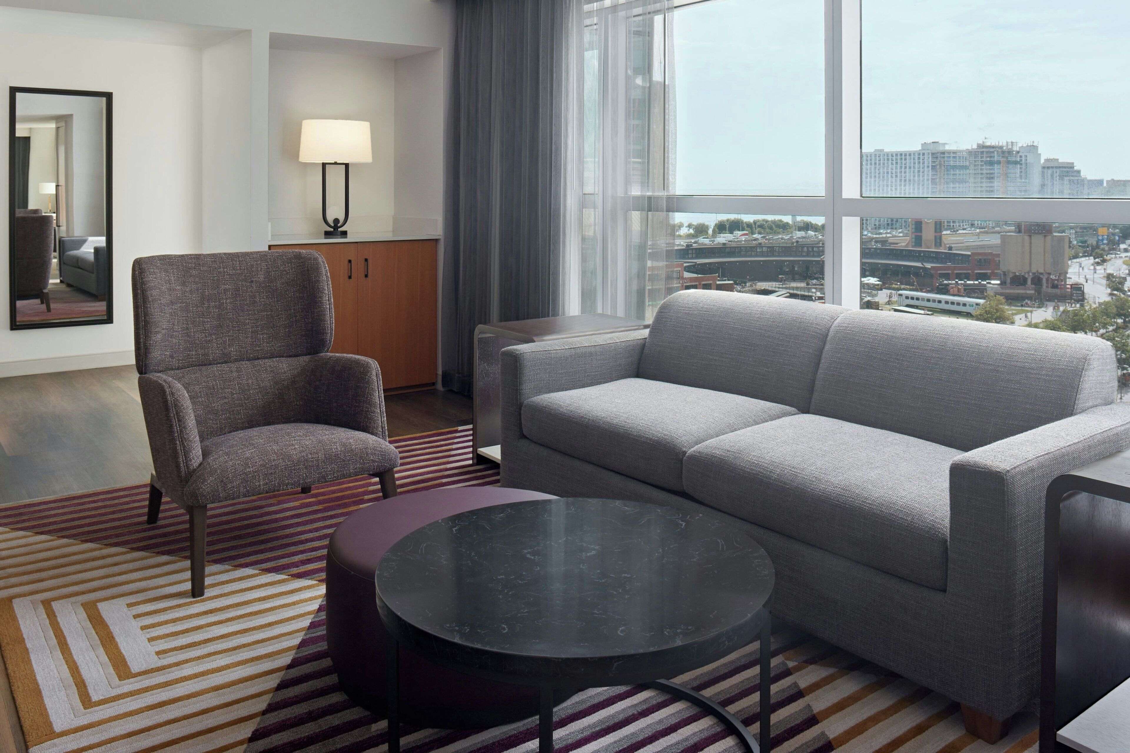 Habitación de hotel moderna con sofá gris, sillón y mesa redonda. Una lámpara está sobre una mesita de noche, y un ventanal ofrece vistas a la ciudad.