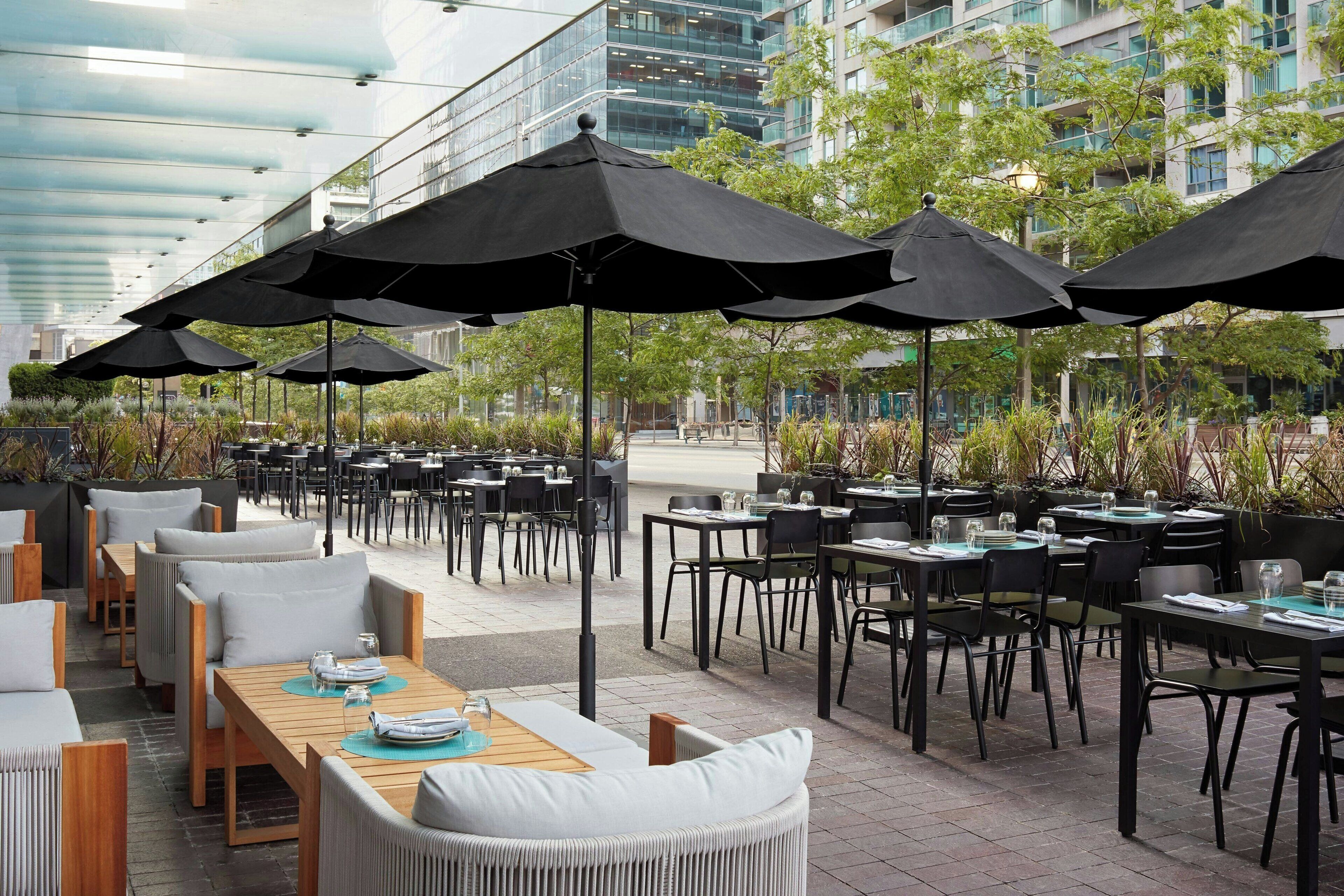 An outdoor dining area with multiple tables and chairs, black umbrellas, and a modern building in the background.