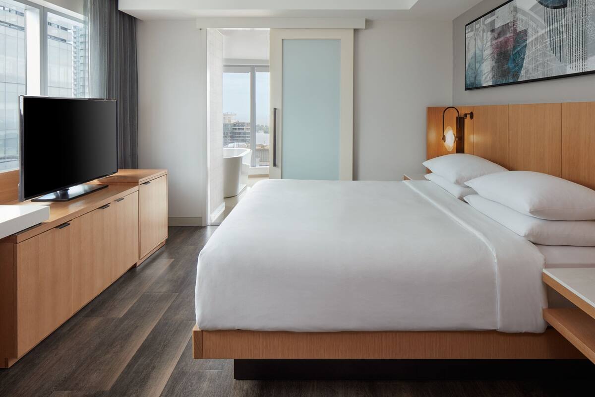 Quarto de hotel moderno com uma cama grande, televisão de tela plana, móveis de madeira e vista para a cidade.