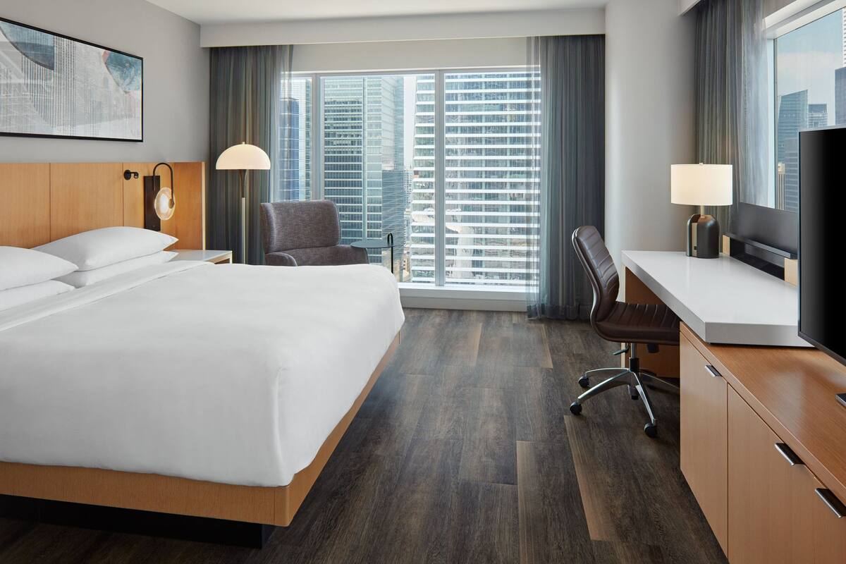 Quarto de hotel com uma cama grande, uma escrivaninha, uma cadeira, uma televisão e uma janela com vista para a cidade.