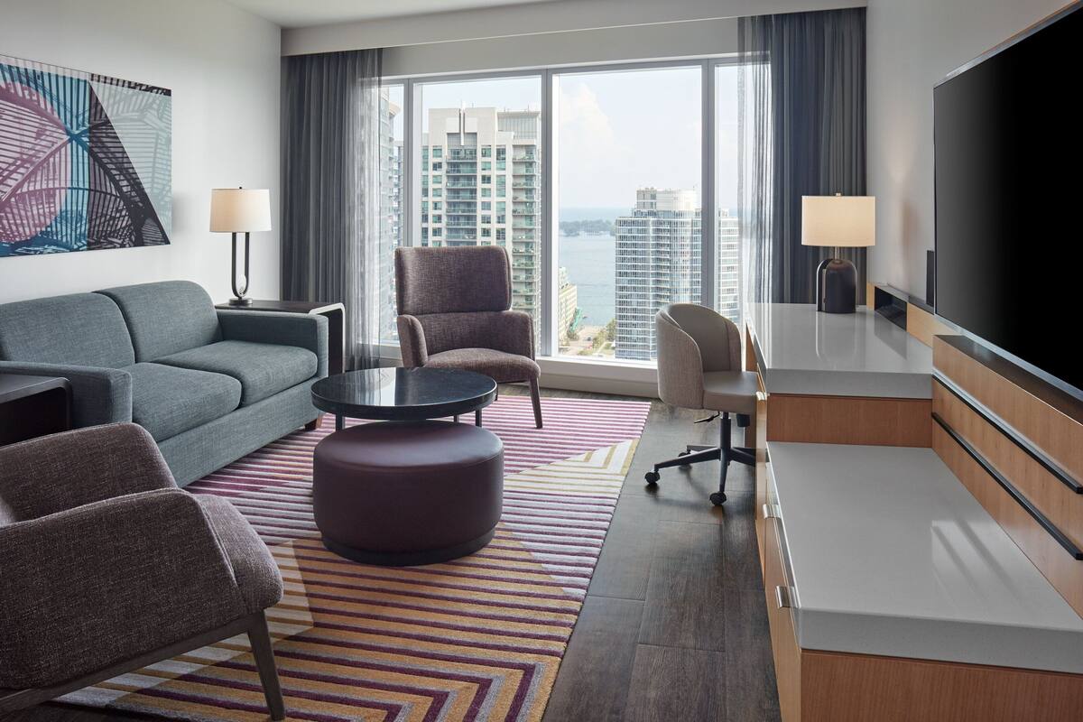 Quarto de hotel moderno com uma grande janela, um sofá, uma cadeira, uma mesinha e uma televisão.