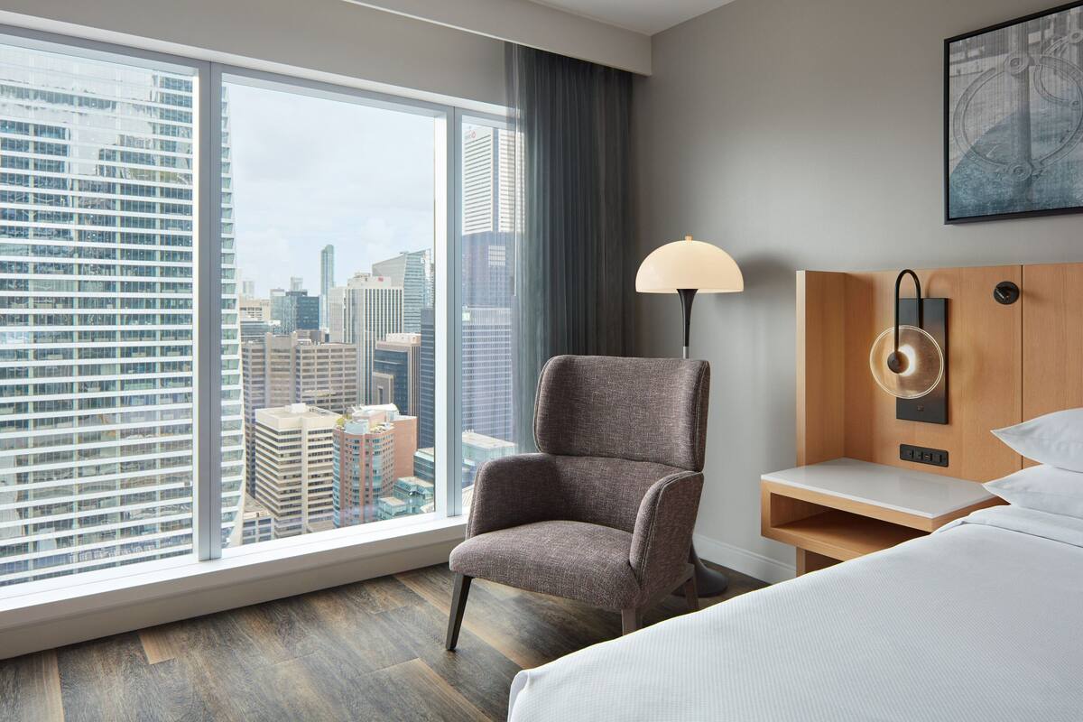 Quarto de hotel com uma janela grande, uma cadeira, mesa de cabeceira e uma cama.