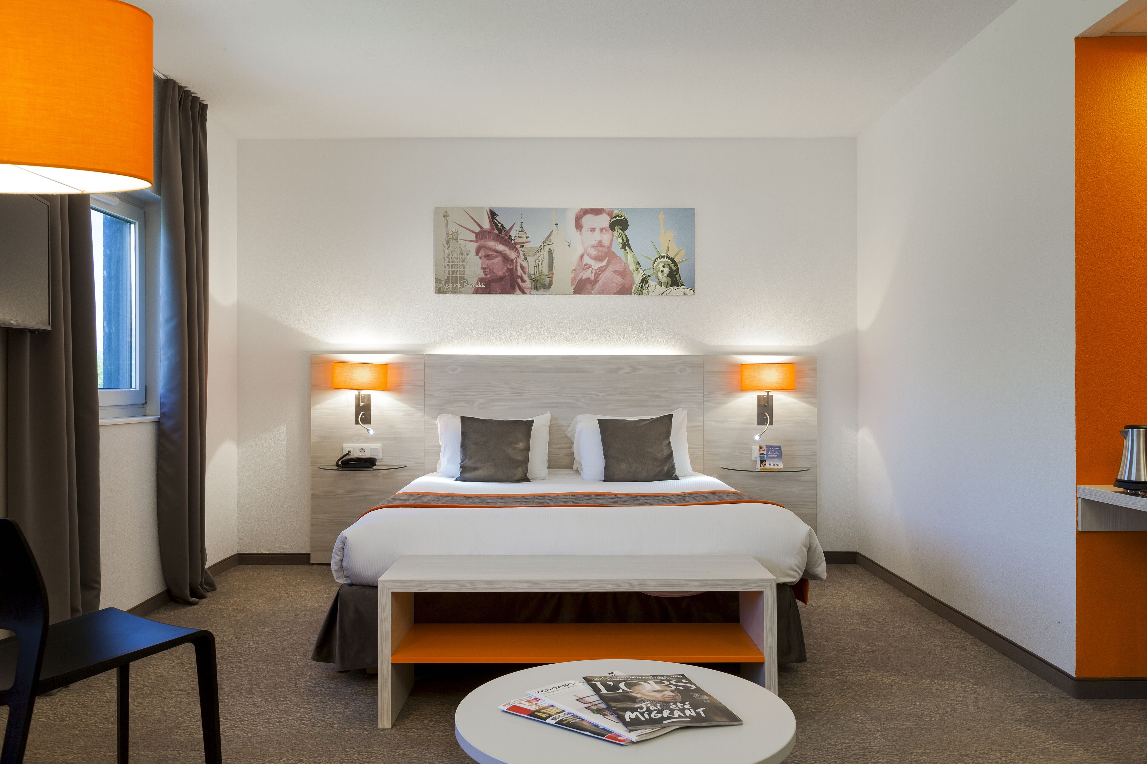 Una habitación de hotel con una cama, lámparas de noche, un escritorio con una silla, una mesa redonda con una revista y un cuadro sobre la cama.