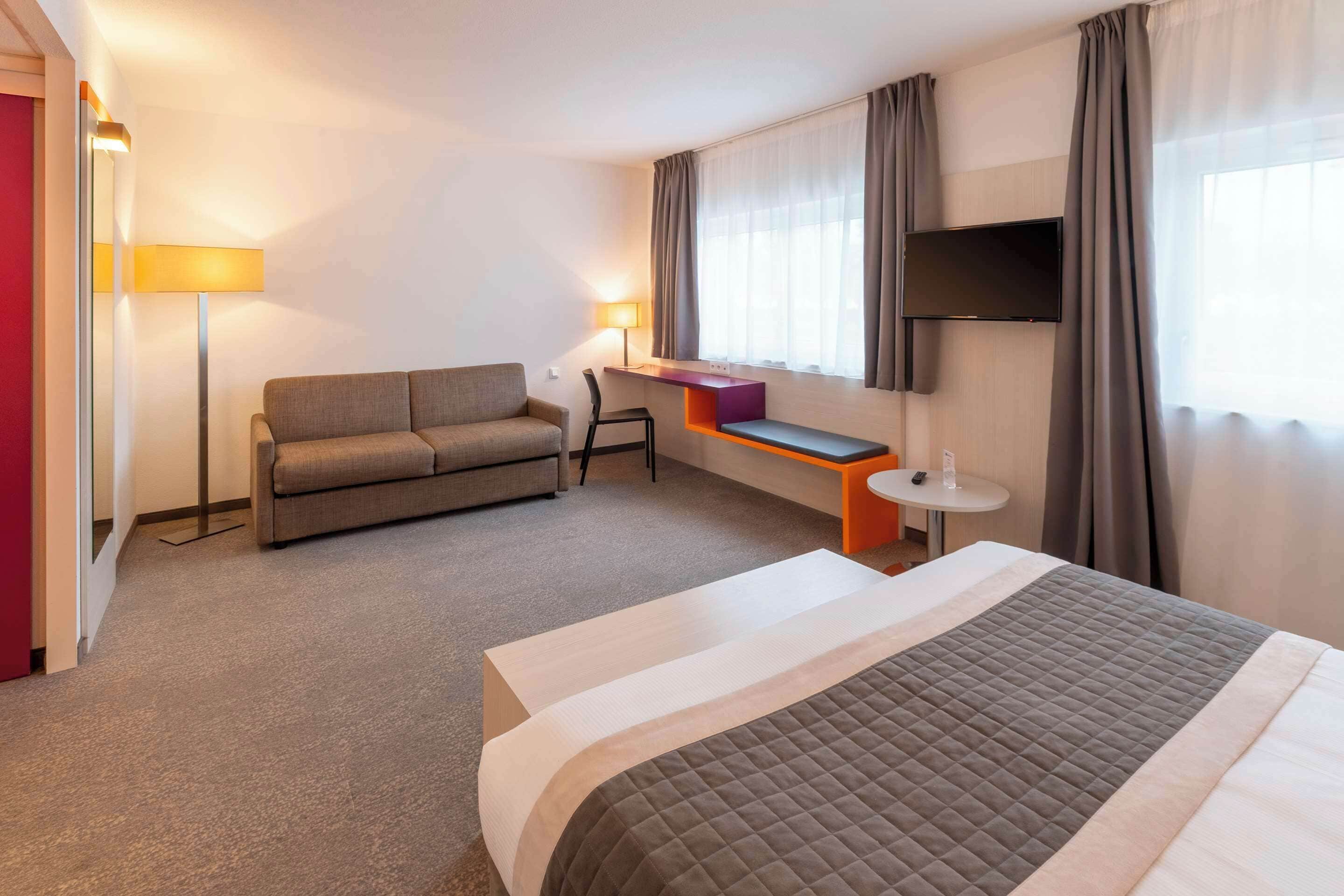 A hotel room with a bed, a sofa, a desk, a TV, and a window with curtains.