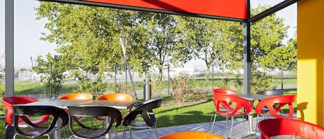 An outdoor dining area with red and black chairs, a table, and a glass enclosure with a view of a green lawn and trees.