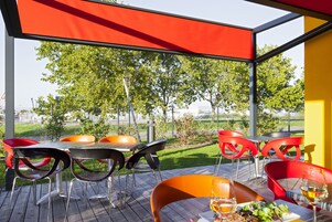 Un área de comedor al aire libre con sillas rojas y negras, una mesa y un cerramiento de cristal con vistas a un césped verde y árboles.