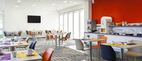 A modern dining area with multiple tables and chairs, a kitchen area with a red accent wall, and a flat-screen TV mounted on the wall.