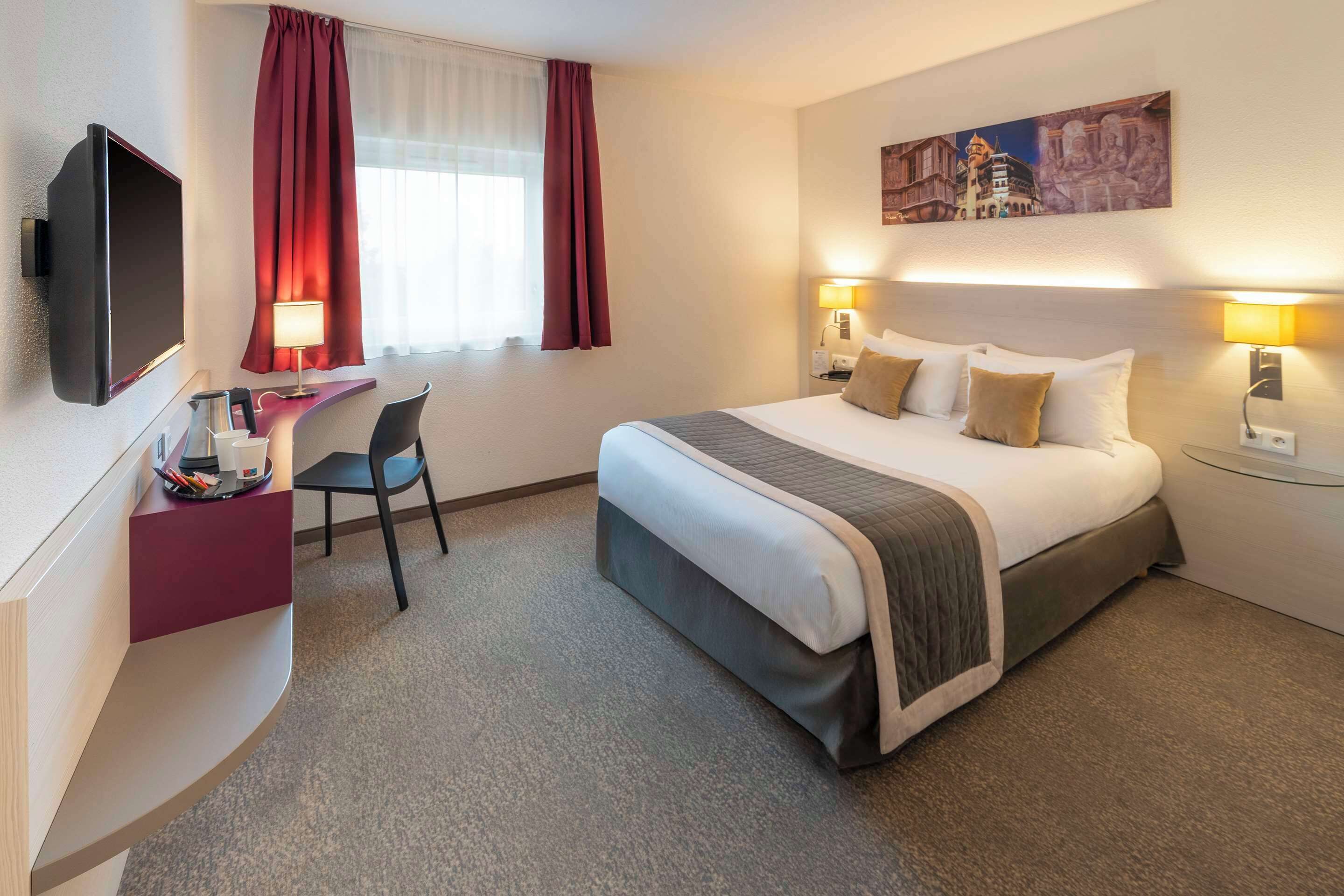 A hotel room with a bed, desk, chair, TV, and artwork on the wall.