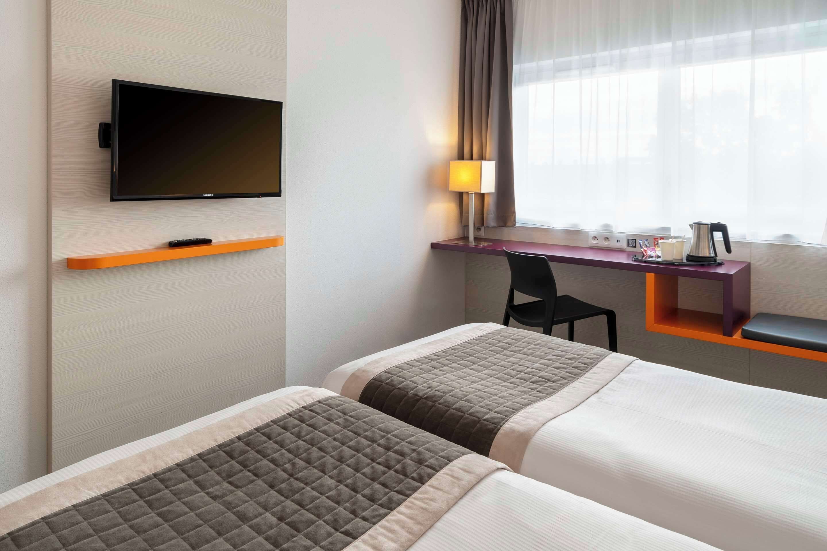 Habitació d'hotel amb dos llits, un televisor fixat a la paret, escriptori amb una cadira, una làmpada i una finestra amb cortines.