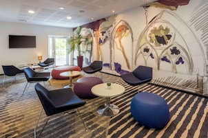 Un área de descanso moderna con asientos negros y azules, una mesa redonda y un mural con diseños florales y arquitectónicos.