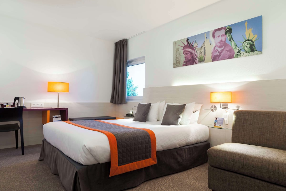 Quarto de hotel com cama, escrivaninha, cadeira e pintura da Estátua da Liberdade.