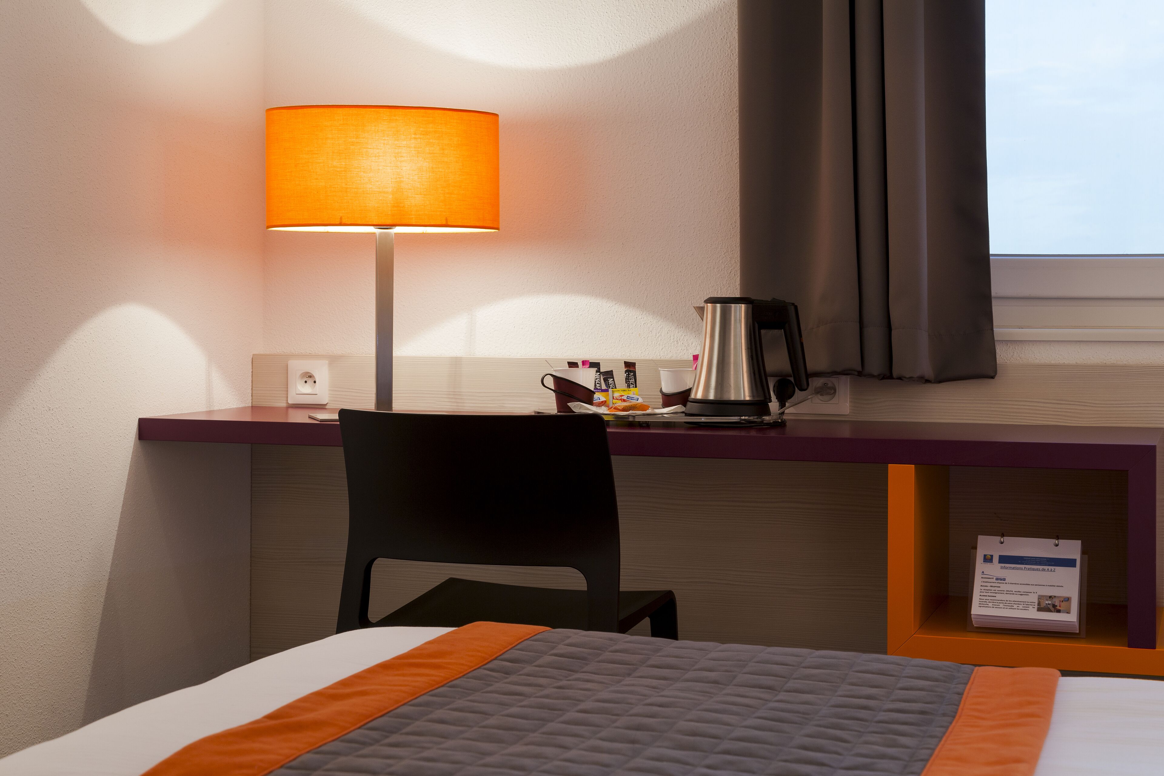 A hotel room with a bed, a desk, a chair, a lamp, a coffee maker, and a window with curtains.