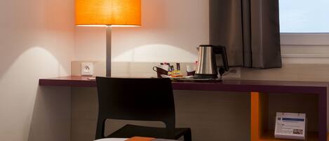A hotel room with a bed, a desk, a chair, a lamp, a coffee maker, and a window with curtains.