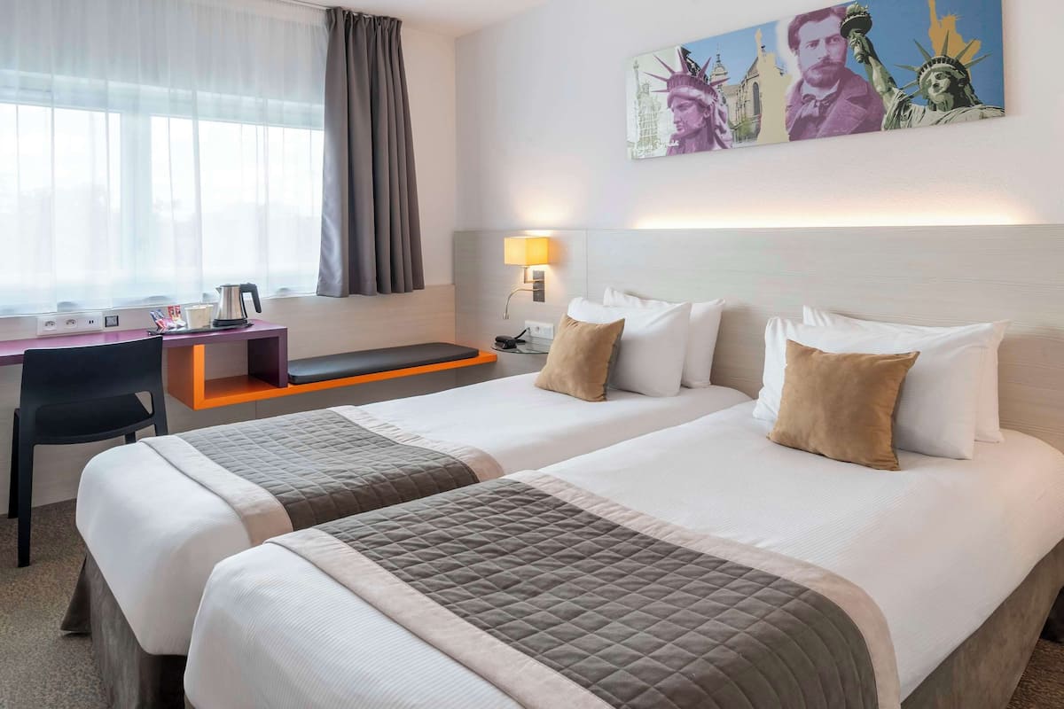 Quarto de hotel com duas camas, uma escrivaninha, uma cadeira, um abajur e um quadro da Estátua da Liberdade.