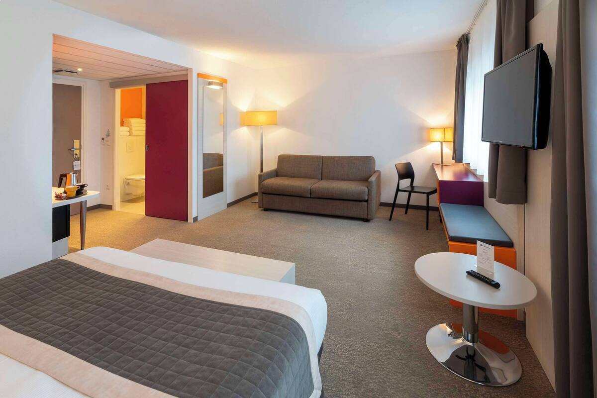 Quarto de hotel com cama, sofá, mesinha, televisão e secretária com abajur.
