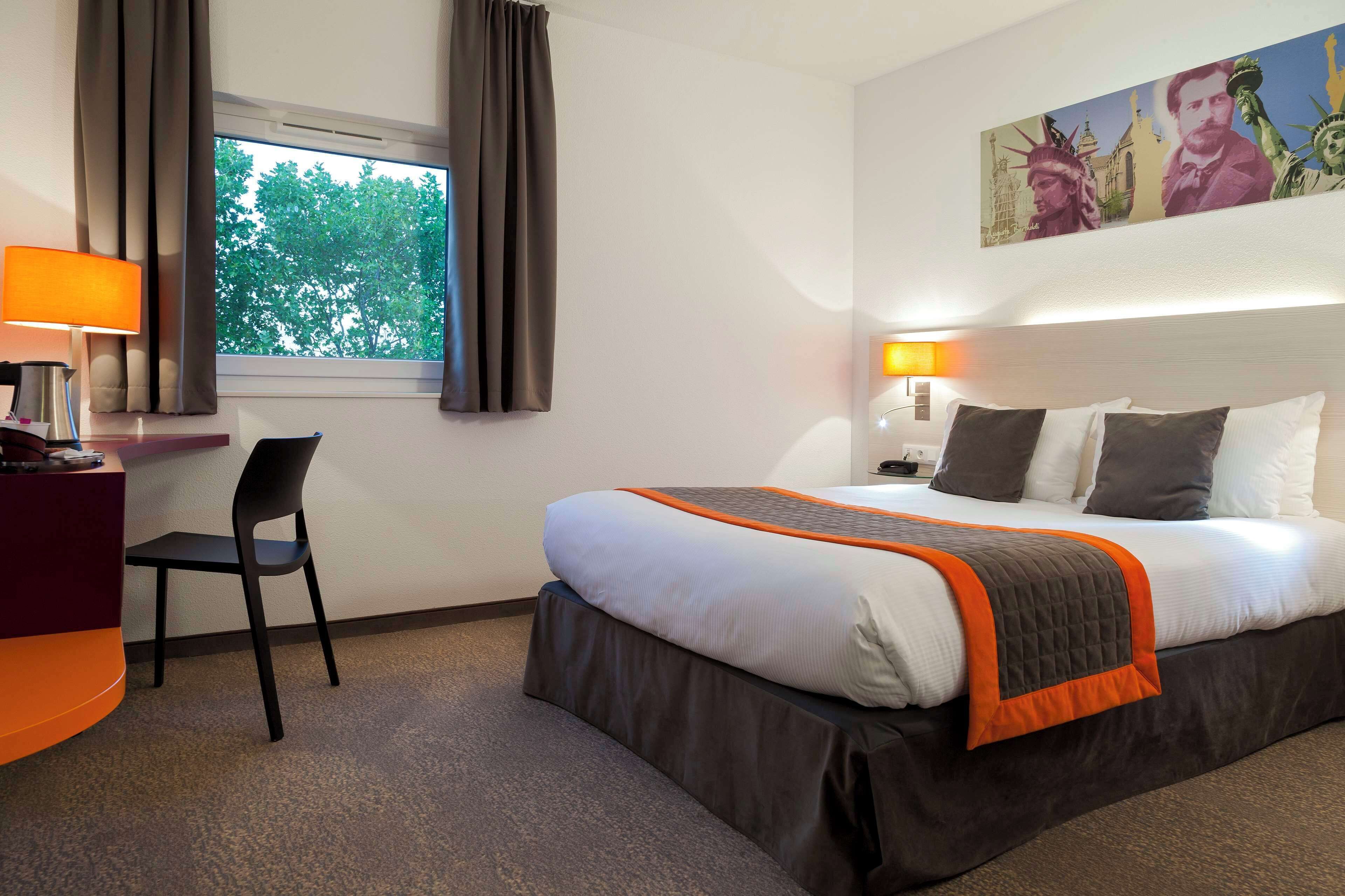 A hotel room with a bed, a desk, a chair, a window with curtains, and a painting on the wall.