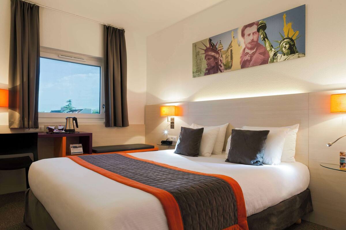 Quarto de hotel com uma cama grande, uma escrivaninha, uma cadeira, uma janela com cortinas, um quadro da Estátua da Liberdade e uma mesa de cabeceira com abajur.