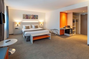 Una habitación de hotel moderna con una cama grande, mesitas de noche con lámparas, un escritorio pequeño con una silla y un armario con estantes.
