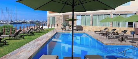 Une piscine avec des chaises longues et des parasols, une marina en arrière-plan et un bâtiment doté de grandes fenêtres.