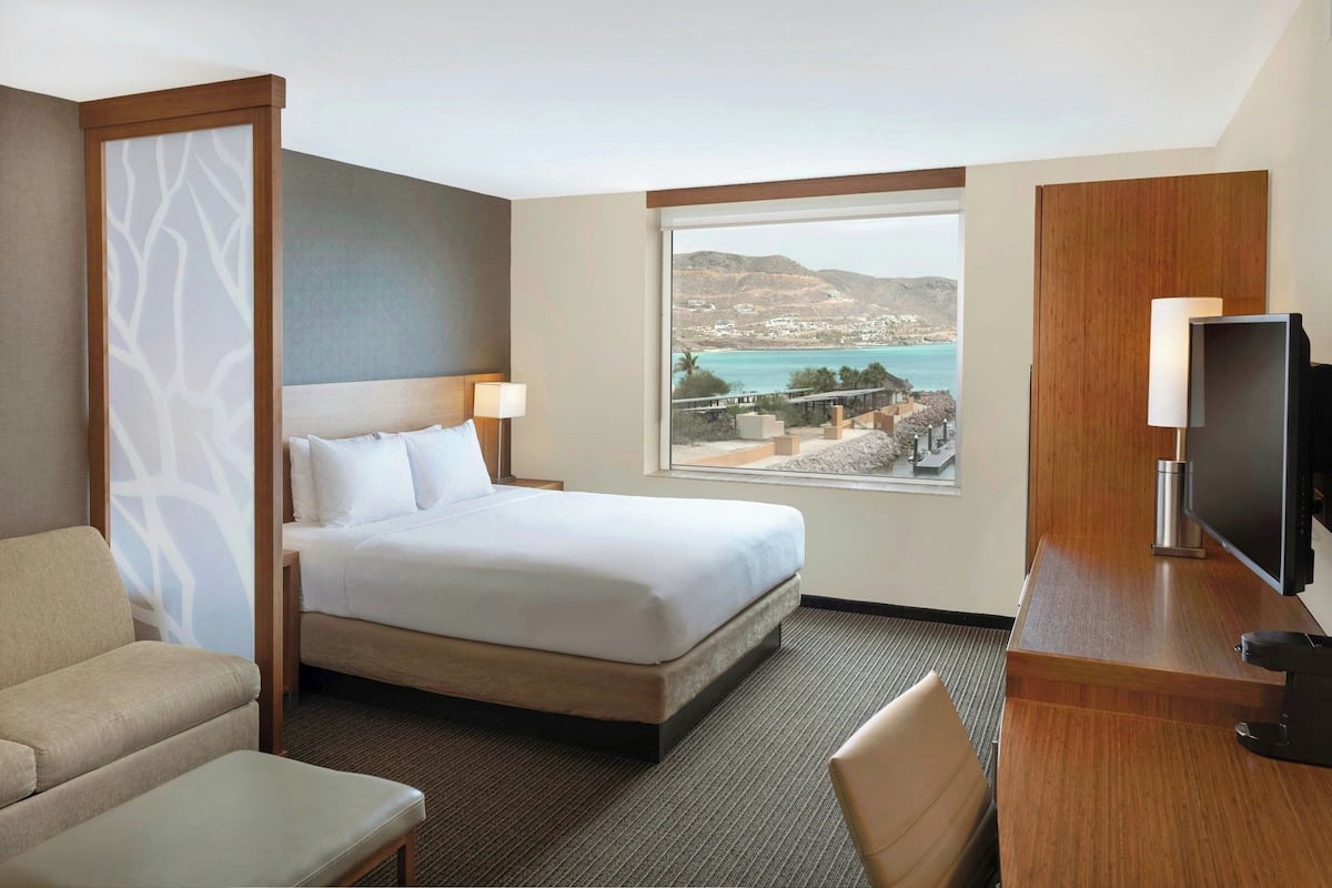 A hotel room with a bed, a desk, a chair, and a large window offering a view of the sea and hills.