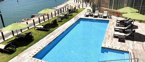 A rectangular swimming pool with a wooden deck surrounding it, equipped with lounge chairs and umbrellas.