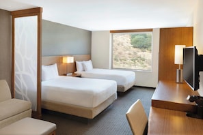 A hotel room with two beds, a desk, a TV, and a view of the outdoors.
