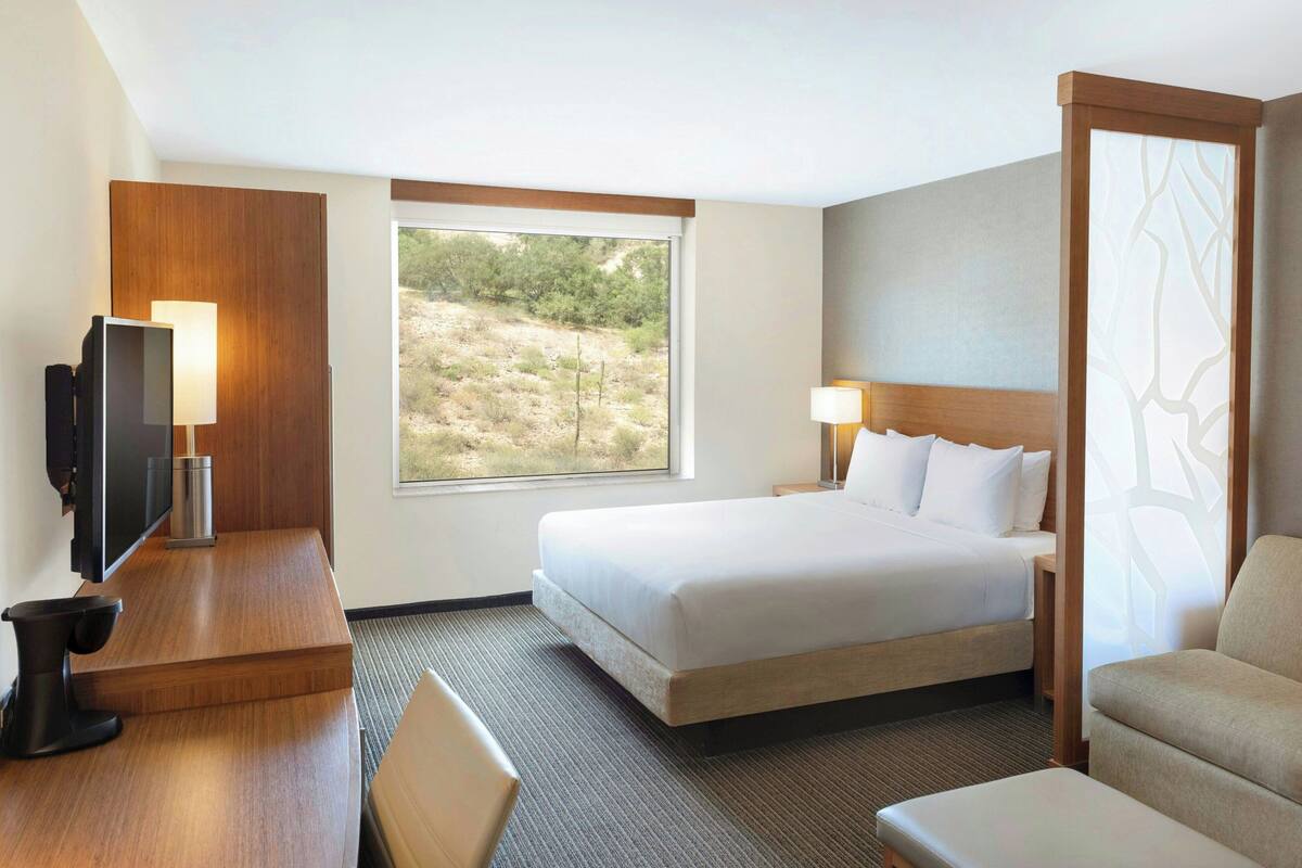 A hotel room with a bed, a desk with a TV, a chair, a sofa, and a large window with a view of a desert landscape.