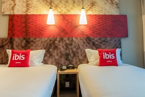Una habitación de hotel con dos camas, cada una con una almohada roja de 'ibis HOTELS', una mesita de noche con teléfono y lámpara, y una pared con textura decorativa.