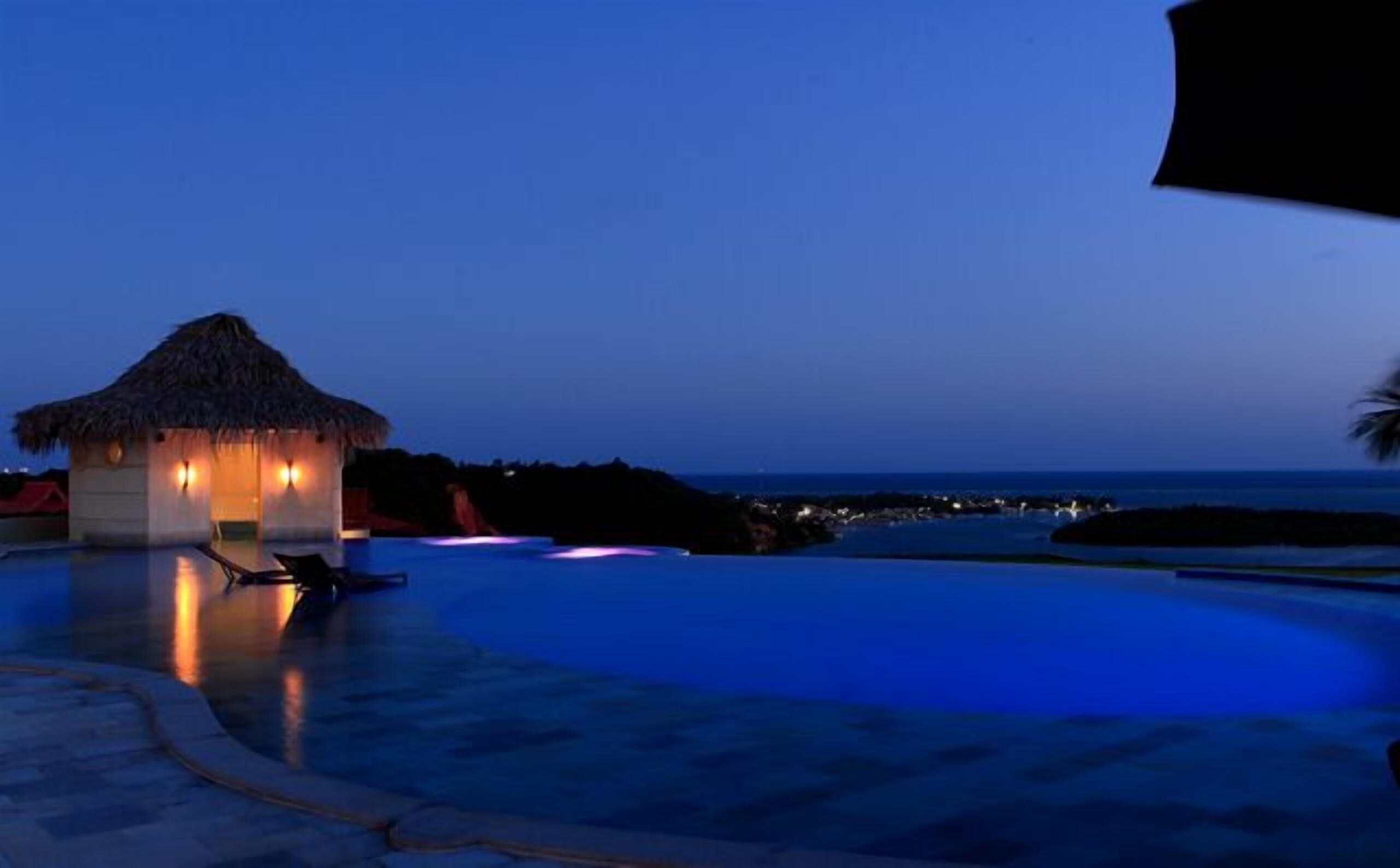Un resort con piscina, una estructura de techo de paja y vista al océano al atardecer.