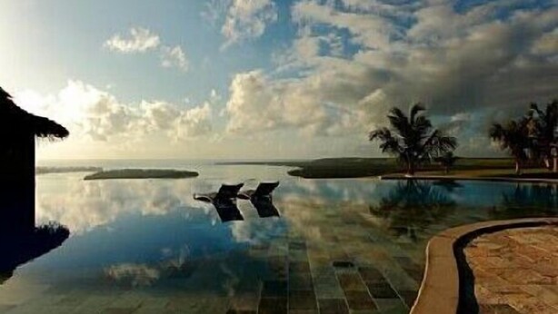 A tranquil infinity pool reflecting the sky and palm trees, surrounded by a paved area and a thatched-roof structure. - Gungaporanga Hotel (Barra de São Miguel)