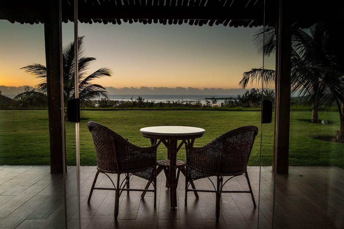 An outdoor seating area with wicker chairs and a table, overlooking a grassy landscape and the ocean at sunset.