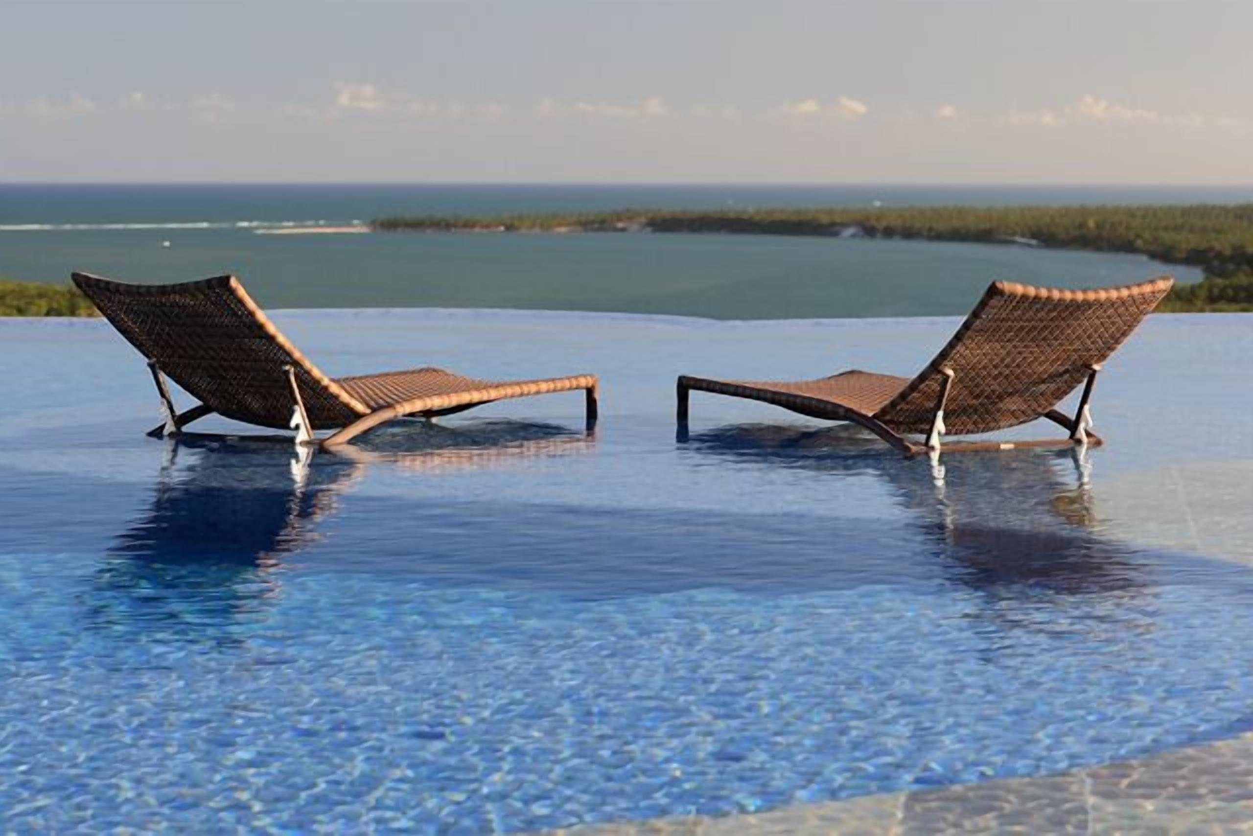 Dos sillones de descanso junto a una piscina infinita de color azul intenso, con vista al océano y una isla distante.
