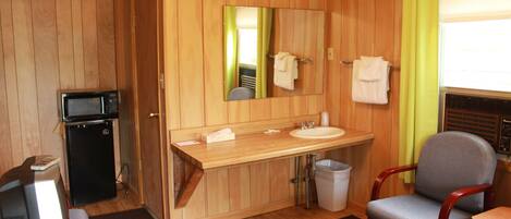 A compact hotel room with a wooden interior, a small kitchenette, a sink, a mirror, a chair, and a window with curtains.