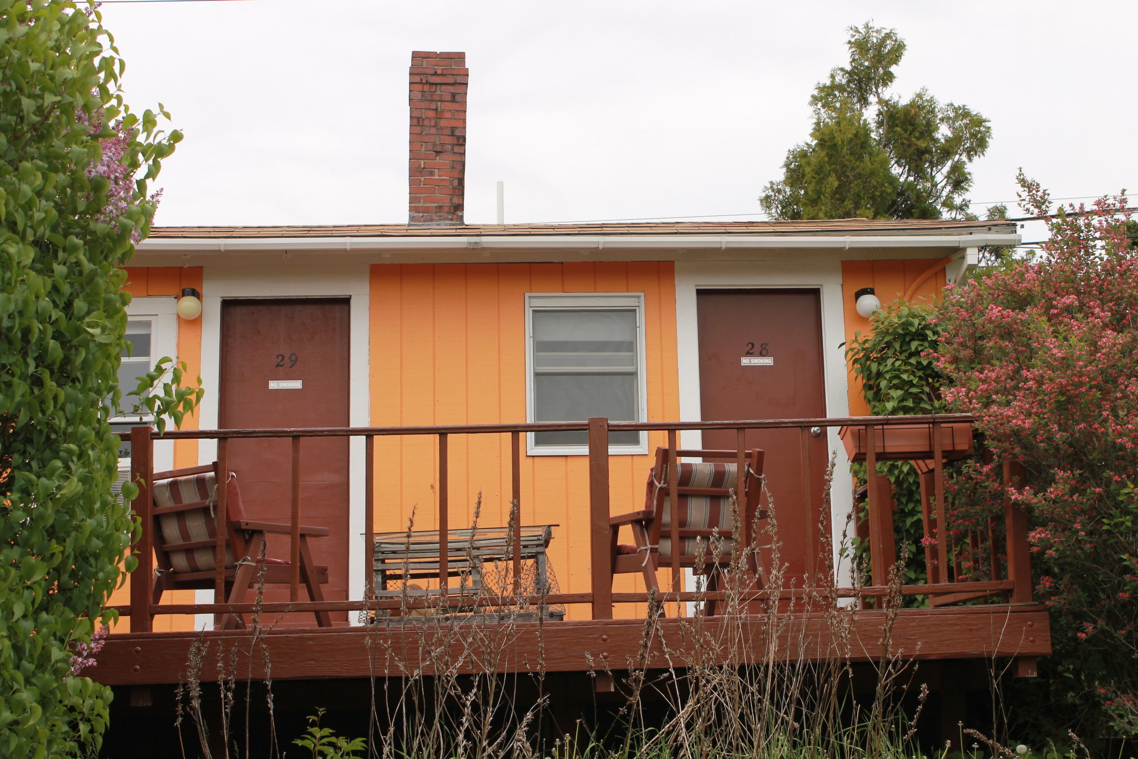 A two-story building with numbered doors, a wooden deck with chairs, and a brick chimney.