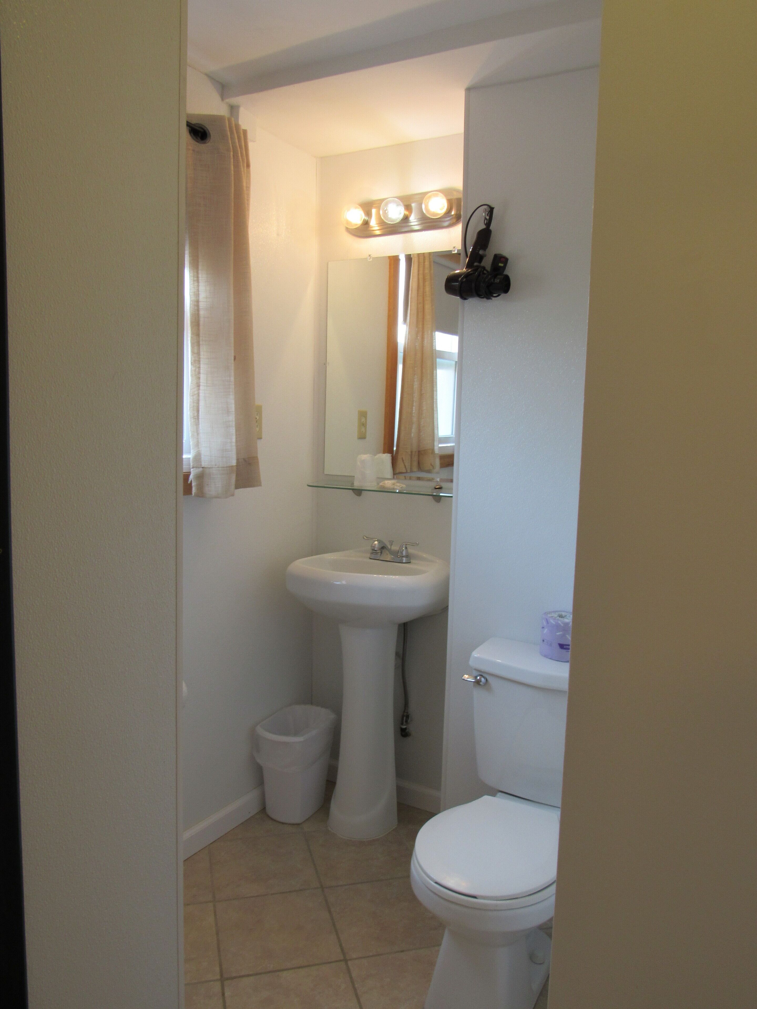 A bathroom with a pedestal sink, a toilet, a mirror with lights, a trash can, and a shower curtain.