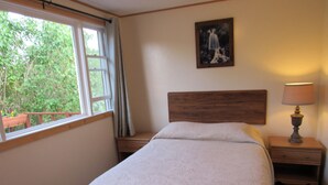 A bedroom with a bed, a nightstand with a lamp, a window with curtains, and a painting on the wall. - Acadia Gateway Motel (Trenton)